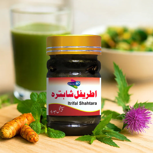 Itrifal Shahtara herbal jar displayed with fresh turmeric, mint leaves, and a thistle flower, with a green juice and salad bowl blurred in the background.