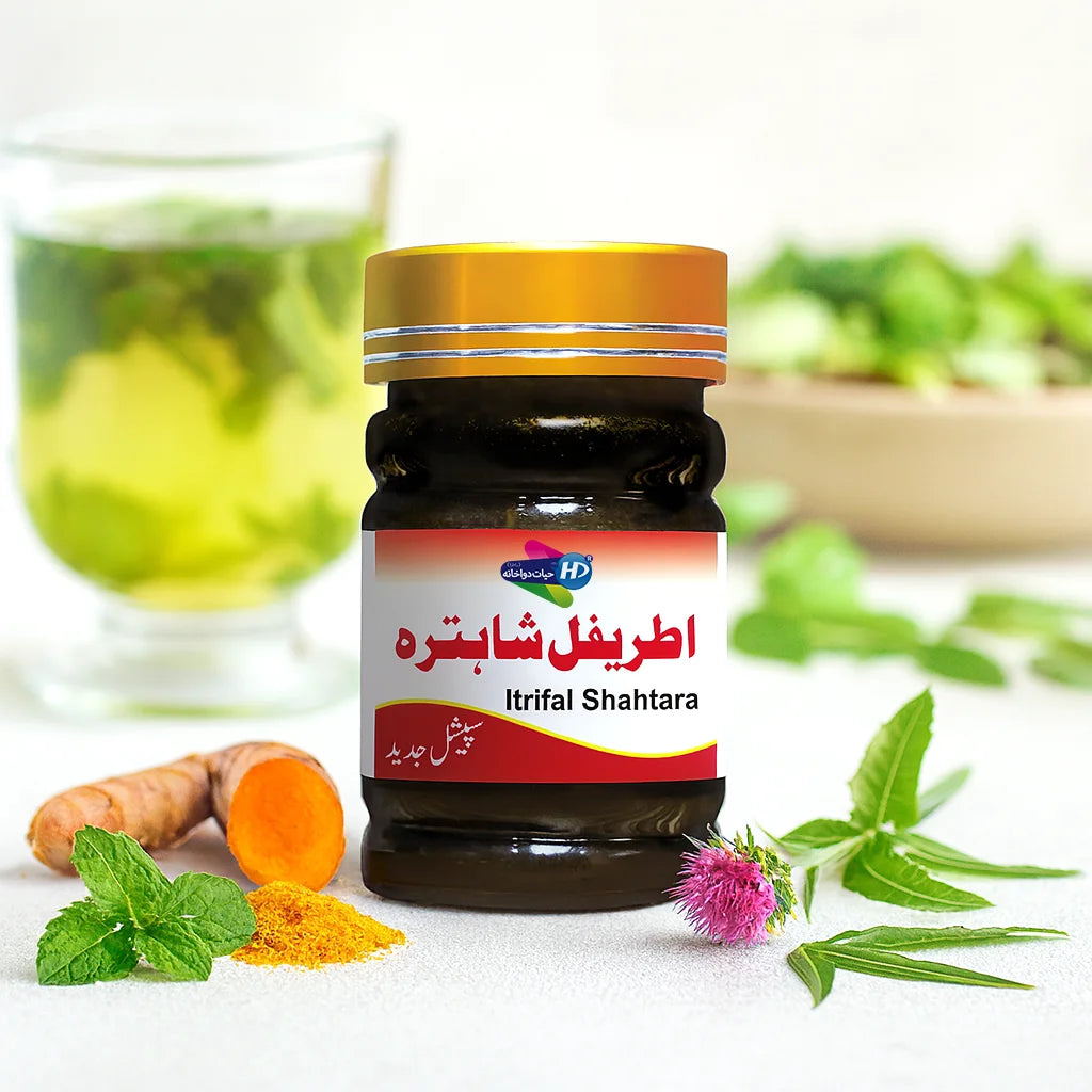 Itrifal Shahtara herbal jar placed on a table with turmeric, mint leaves, and a purple thistle flower beside it, with a glass of green herbal drink in the background.