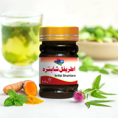 Itrifal Shahtara herbal jar placed on a table with turmeric, mint leaves, and a purple thistle flower beside it, with a glass of green herbal drink in the background.
